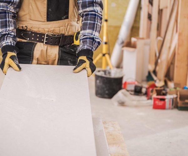 Male Construction Worker Holding Long Piece Of Gray Drywall Material Ready To Be Installed On House Wall.