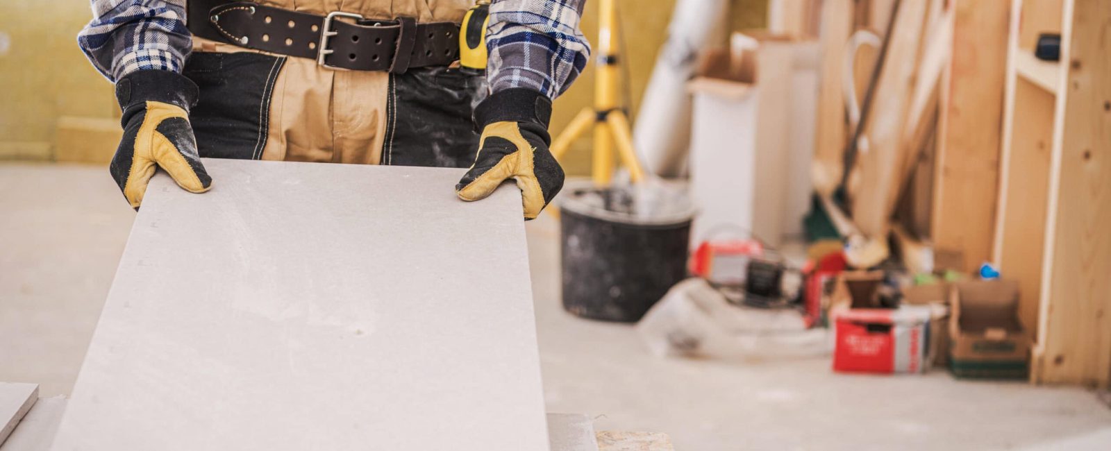 Male Construction Worker Holding Long Piece Of Gray Drywall Material Ready To Be Installed On House Wall.